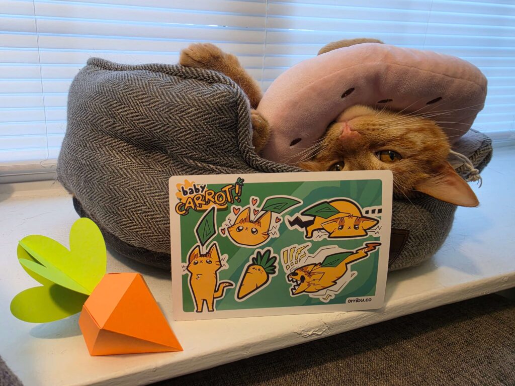 Pictured: a papercraft carrot (left), a sticker sheet featuring charicatures of Carrot (center), and Carrot, an orange tabby, looking at the camera (top).