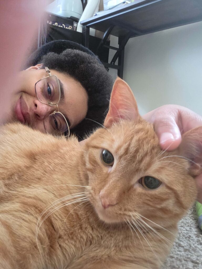 Carrot, an orange tabby cat, looks into the camera. Behind him is Olive, his mom, who's petting him with one hand behind his left ear.